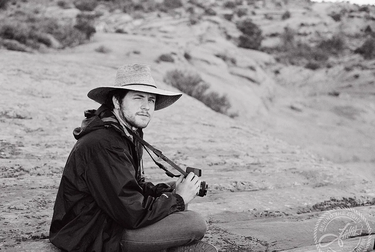 Black-and-white photo of John Matthew Gillen in a wide-brimmed hat, sitting on desert rock with a film camera