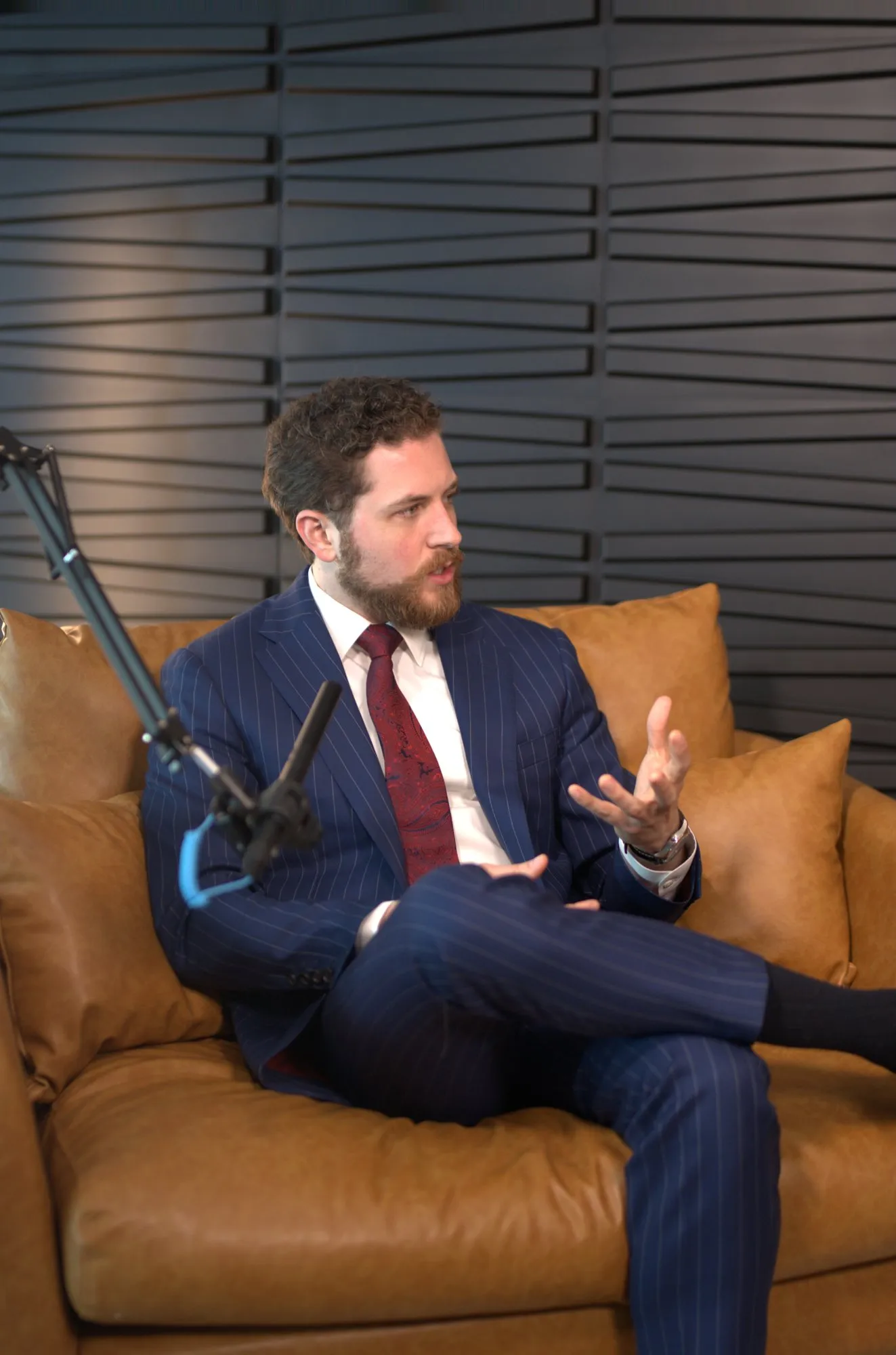 John Matthew Gillen in a navy pinstripe suit and red tie, speaking on a leather couch on the set of The Milk Road Show