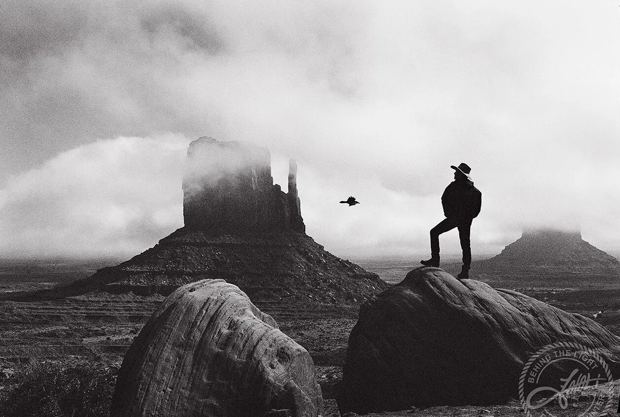 Black-and-white photo of John Matthew Gillen in a cowboy hat standing on a boulder in Monument Valley, a raven in flight beside a fog-covered butte